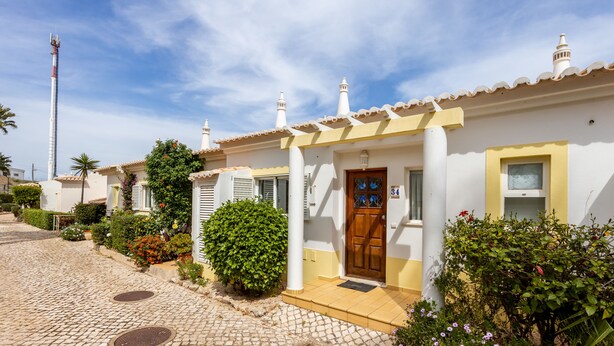Un edificio blanco con puerta de madera y ventana, rodeado de vegetación y pavimento de adoquines.