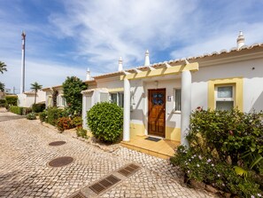 A white building with a wooden door and a window, surrounded by greenery and cobblestone pavement. - Townhouse da Amarela (Budens)