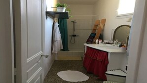 Un baño estrecho con ducha, lavabo y espejo. Hay una toalla colgada en la pared y una pequeña planta en un estante.