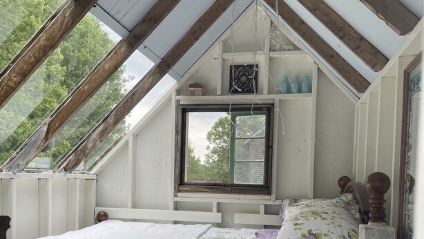 Une chambre avec un plafond en pente, un lit recouvert d’une literie à motifs floraux, une fenêtre et une vue sur les arbres.