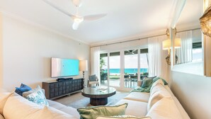 A spacious living room with a large sofa, a coffee table, and a TV. There is a balcony with a view of the ocean. - Beachcomber 3 by Grand Cayman Villas (7 Mile Beach)