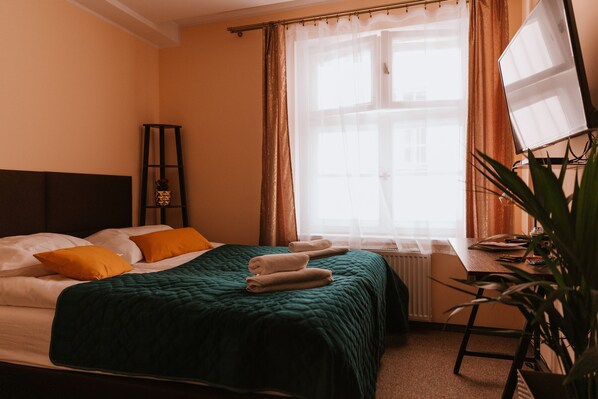 Un lit bien fait, recouvert d’une couette verte, avec deux oreillers et deux serviettes pliées. On y trouve une petite table sur laquelle sont posés une plante et une lampe.