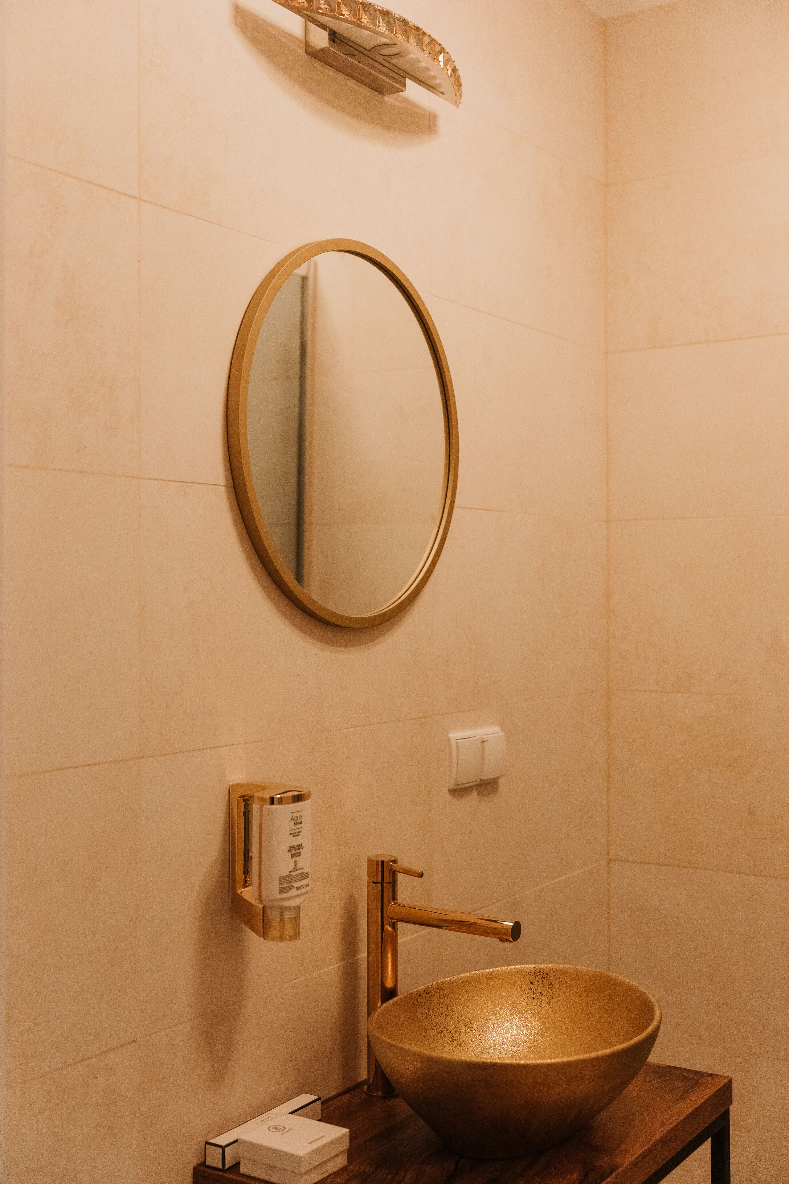 Une salle de bain avec un miroir rond, un lavabo doré et un distributeur de savon fixé au mur.