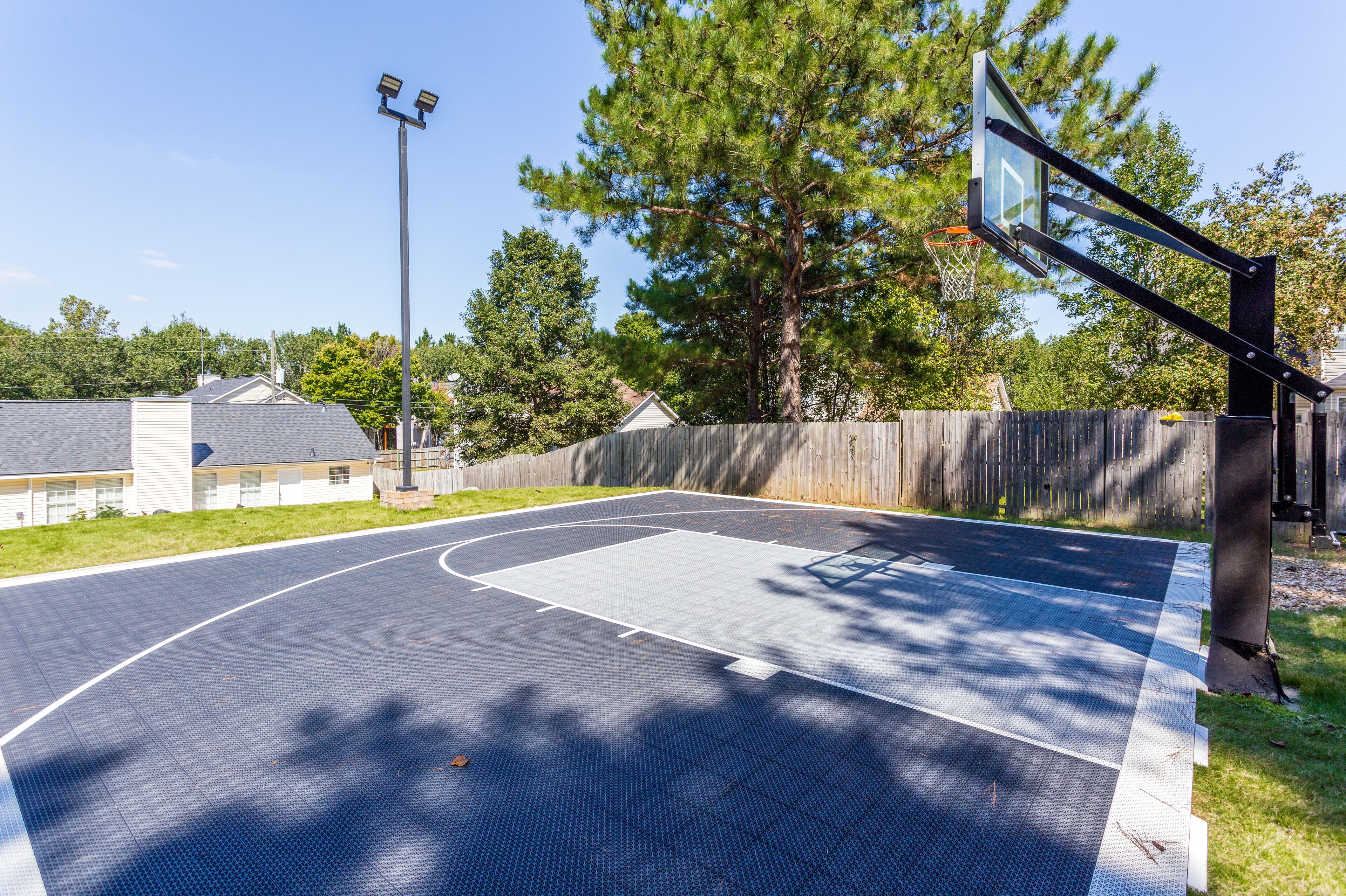 An outdoor basketball court with a wooden fence, trees, and a building in the background.