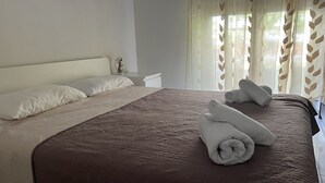A neatly made bed with a brown bedspread and white pillows, a small bedside table with a vase, and sheer curtains with leaf patterns. - Vero Resort Townhouse Hanioti (Халкидики)