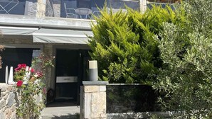 A two-story building with a stone facade, a covered patio with outdoor furniture, and a large evergreen shrub in the foreground. - Vero Resort Townhouse Hanioti (Халкидики)