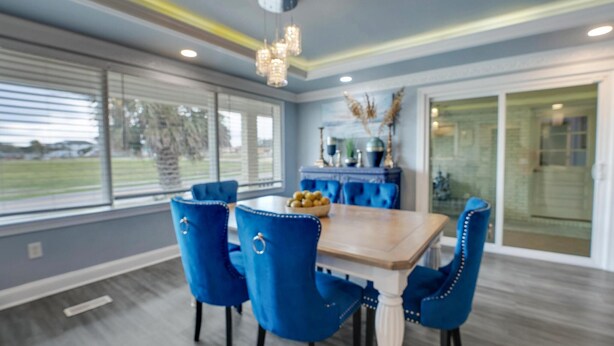 A dining area with a wooden table and blue upholstered chairs, a chandelier, and a view of the outdoors through large windows.