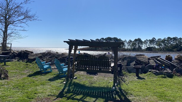 A wooden pergola with a bench, surrounded by a grassy area with blue chairs, overlooking a body of water.