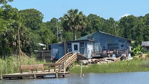 A wooden dock leading to a small house with a porch, surrounded by water and greenery.