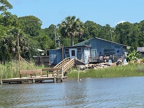 A wooden dock leading to a small house with a porch, surrounded by water and greenery.