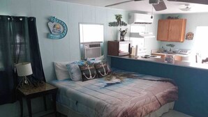 A bedroom with a bed, a nightstand, a window with curtains, a wall clock, and a small table with a lamp.