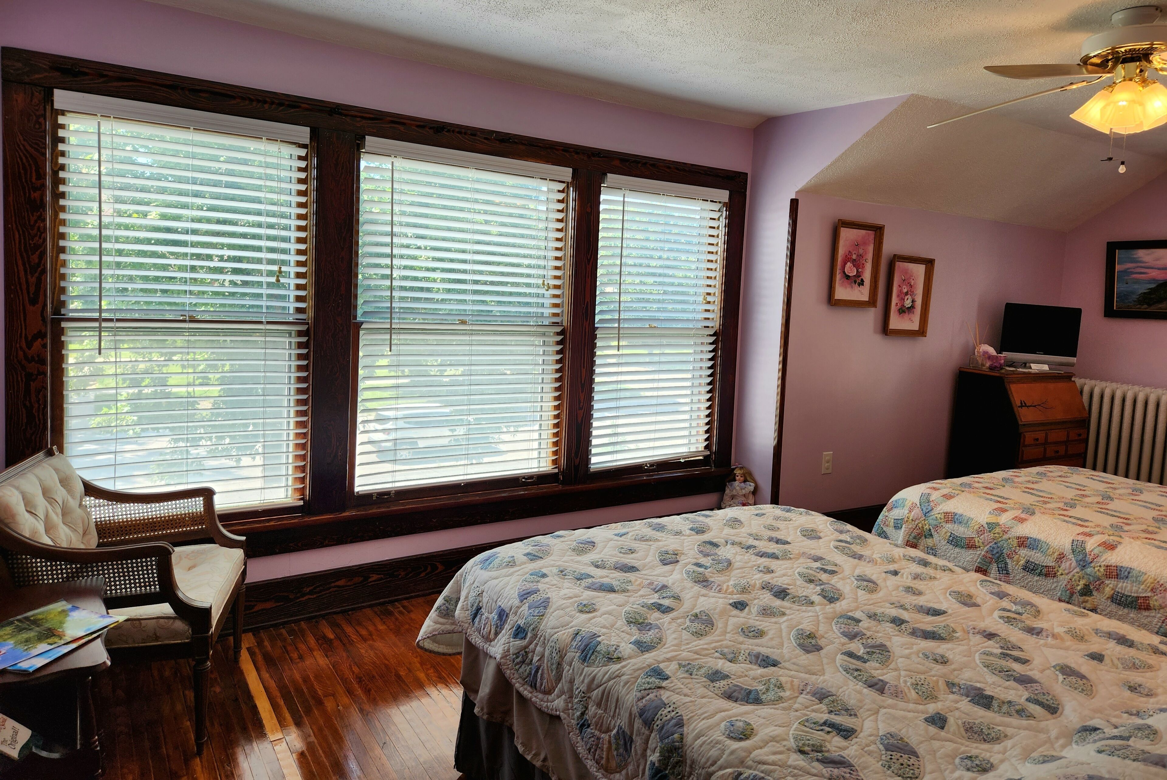 Quarto com duas camas, uma cadeira, escrivaninha e janelas amplas com persianas.