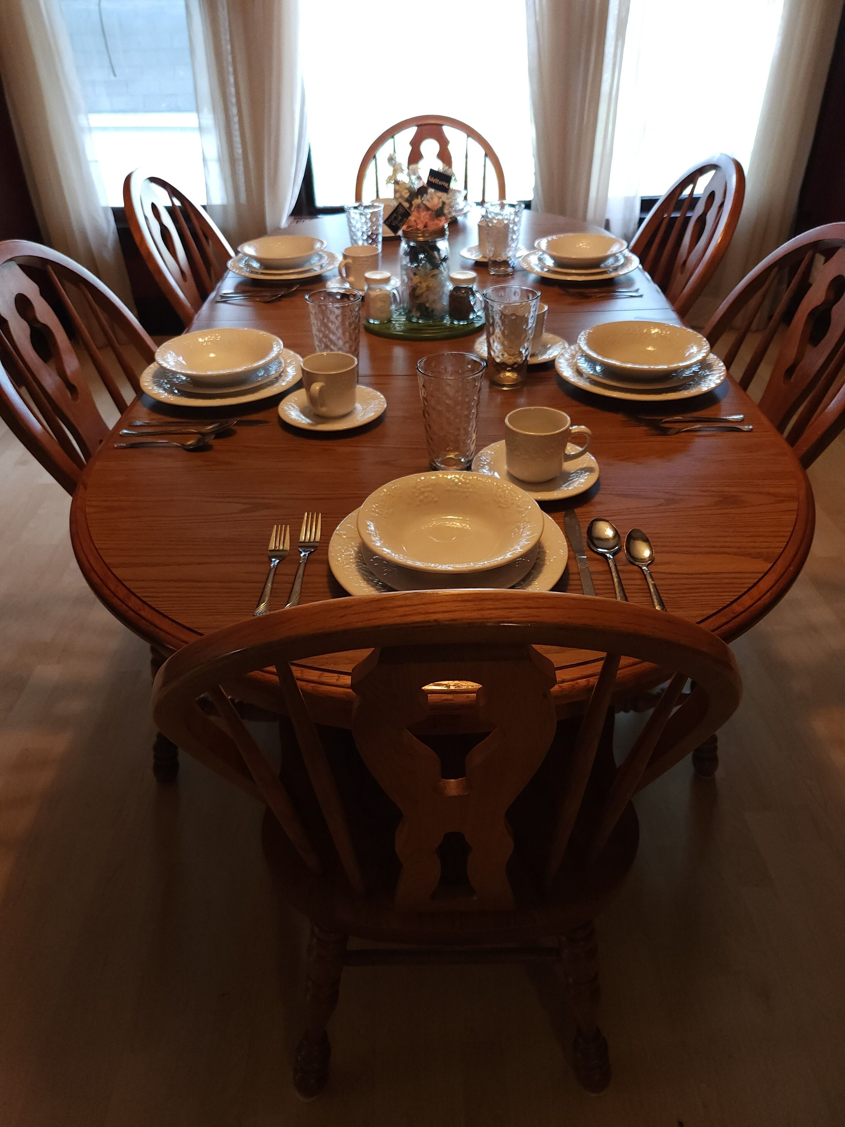 Une table à manger dressée pour six personnes, avec de l’argenterie blanche, des assiettes, des bols et des verres, entourée de chaises en bois.