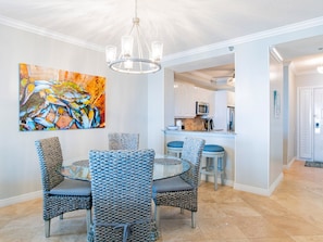 A dining area with a glass table and woven chairs, a colorful abstract painting on the wall, and a kitchen in the background.