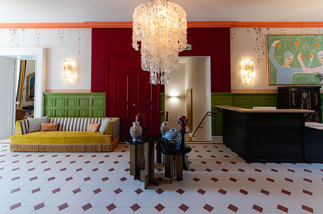 A lobby with a patterned floor, a sofa, a table with decorative items, a chandelier, a reception desk, and a painting on the wall.