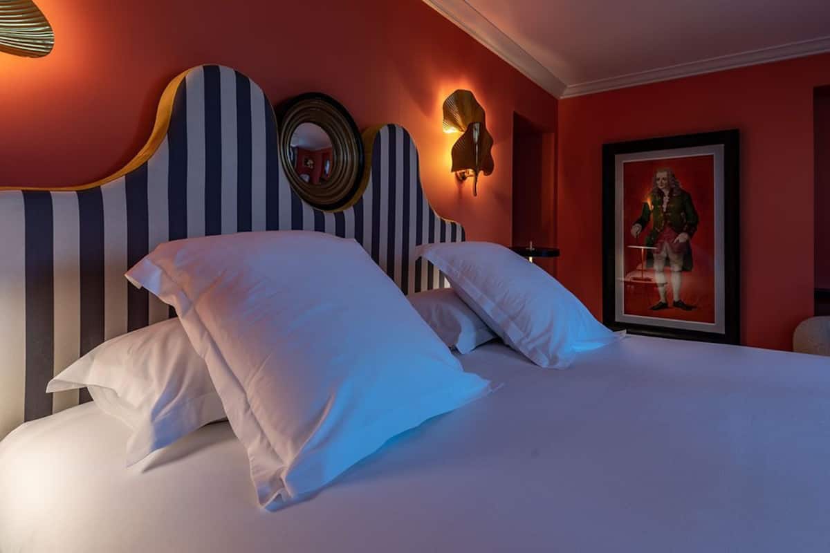 Une chambre d’hôtel avec un grand lit, une tête de lit à rayures, un miroir, des appliques murales et un portrait encadré.