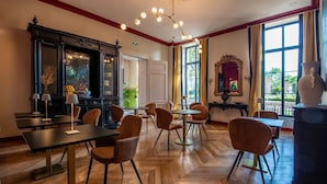 Uma área de refeições com piso de madeira, várias mesas e cadeiras, um lustre decorativo e grandes janelas com cortinas.