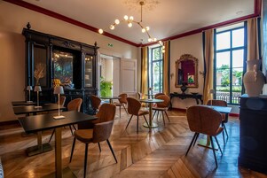 Un comedor con suelo de madera, varias mesas y sillas, una lámpara decorativa y ventanas grandes con cortinas.