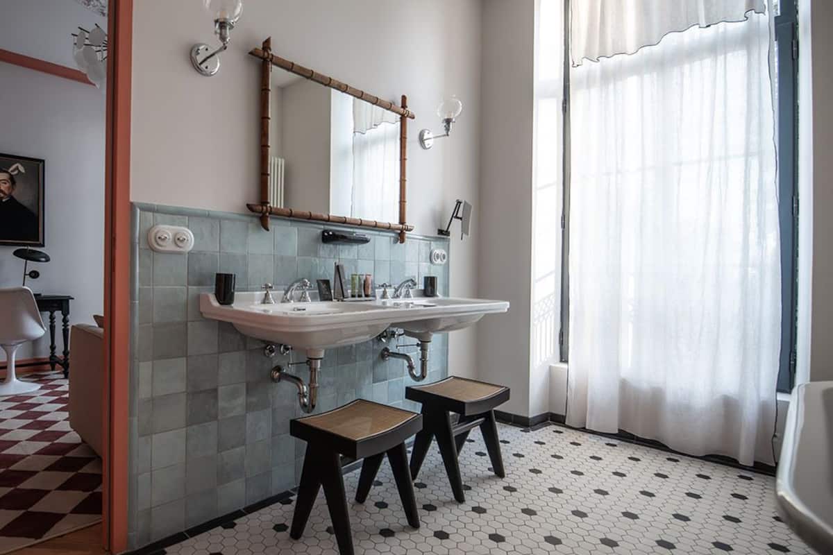 Une salle de bain avec un double vasque, deux tabourets, un miroir et une fenêtre ornée de rideaux légers.
