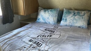 A bed with a blanket featuring names and dates, wooden cabinets above, and a window with a curtain.
