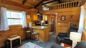 Un acogedor interior de cabaña con techo de madera, una zona de cocina, un espacio de descanso con sofá y sillones, y una alfombra estampada.