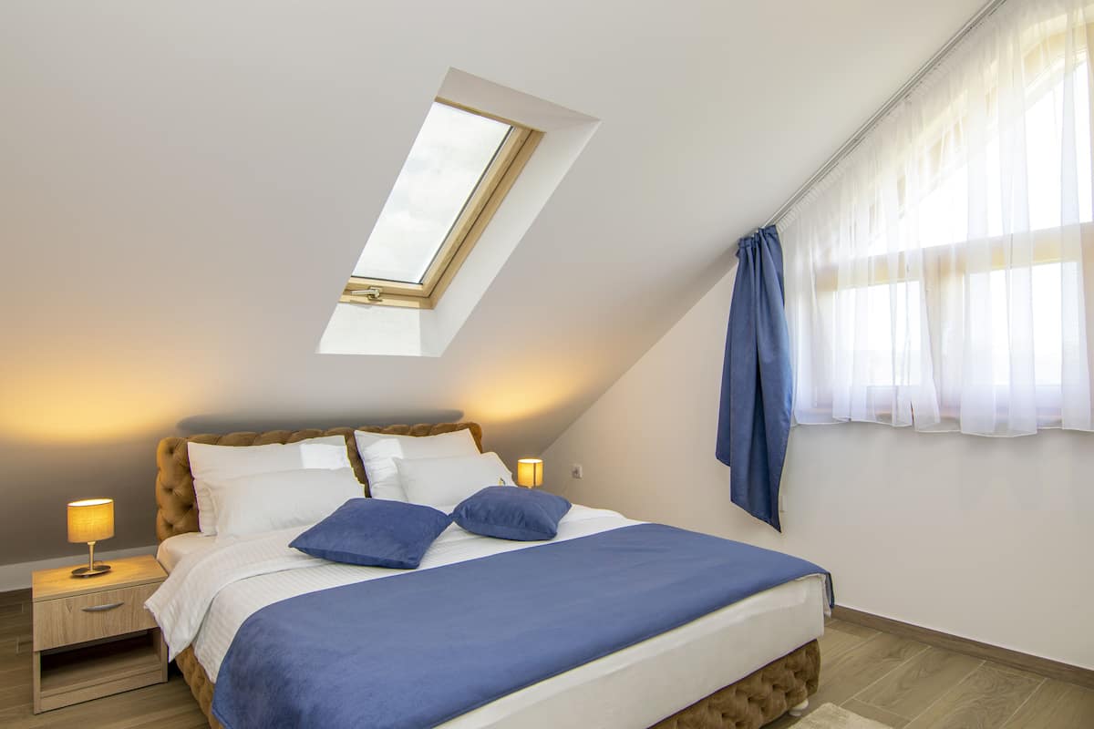A bedroom with a sloped ceiling, a skylight, a bed with blue bedding, a nightstand with a lamp, and a window with sheer curtains.