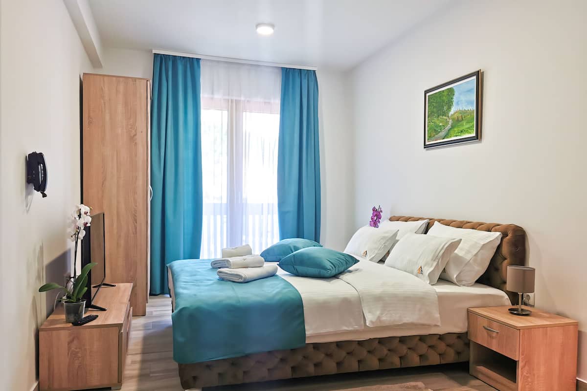 A neatly arranged bedroom with a bed, bedside tables, a nightstand, a potted plant, and a painting on the wall.