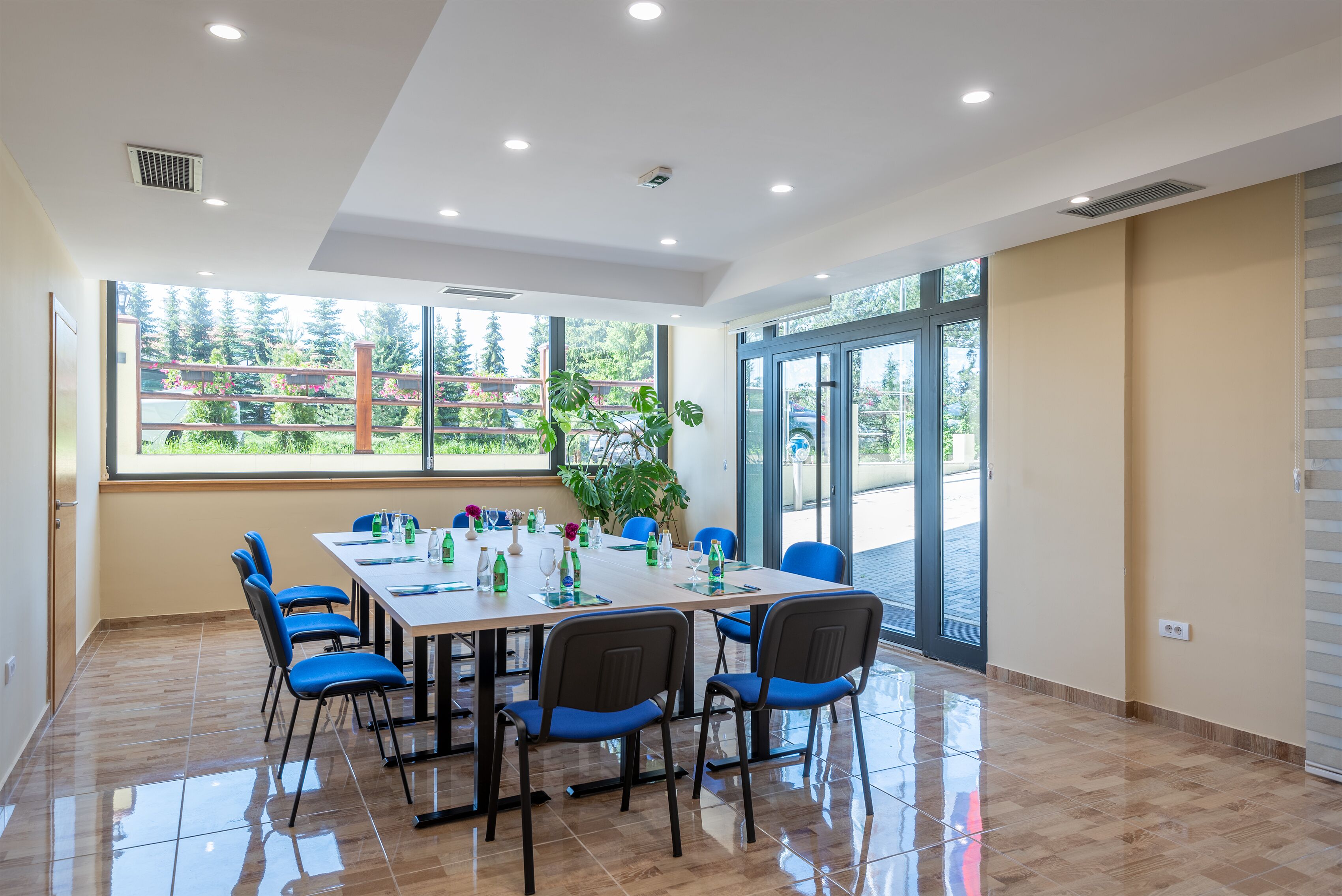A modern conference room with a large table, blue chairs, and large windows offering a view of greenery outside.