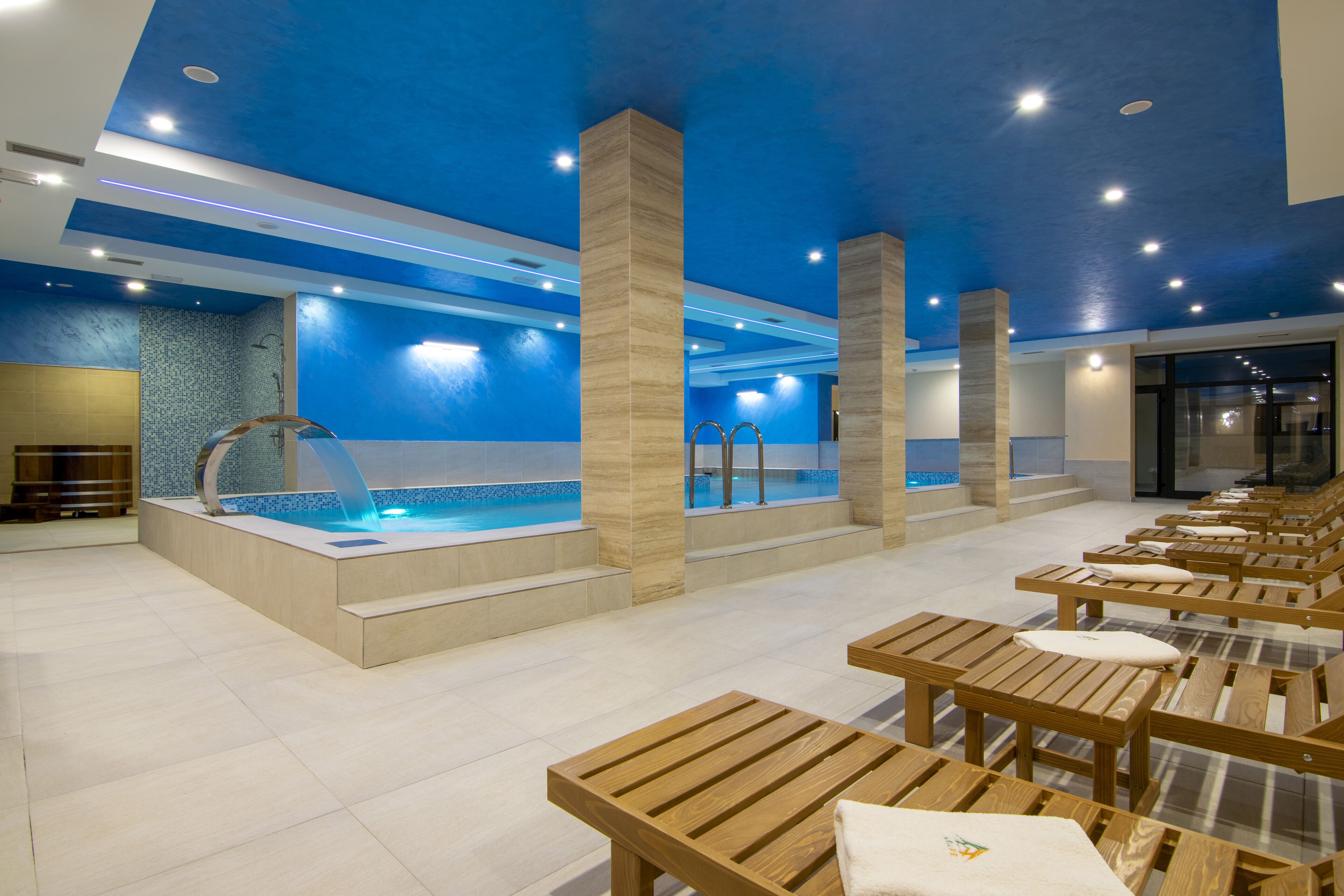 A modern indoor pool area with a blue ceiling, wooden benches, and a central pool with a blue mosaic border.