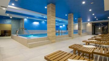 A modern indoor pool area with a blue ceiling, wooden benches, and a central pool with a blue mosaic border.