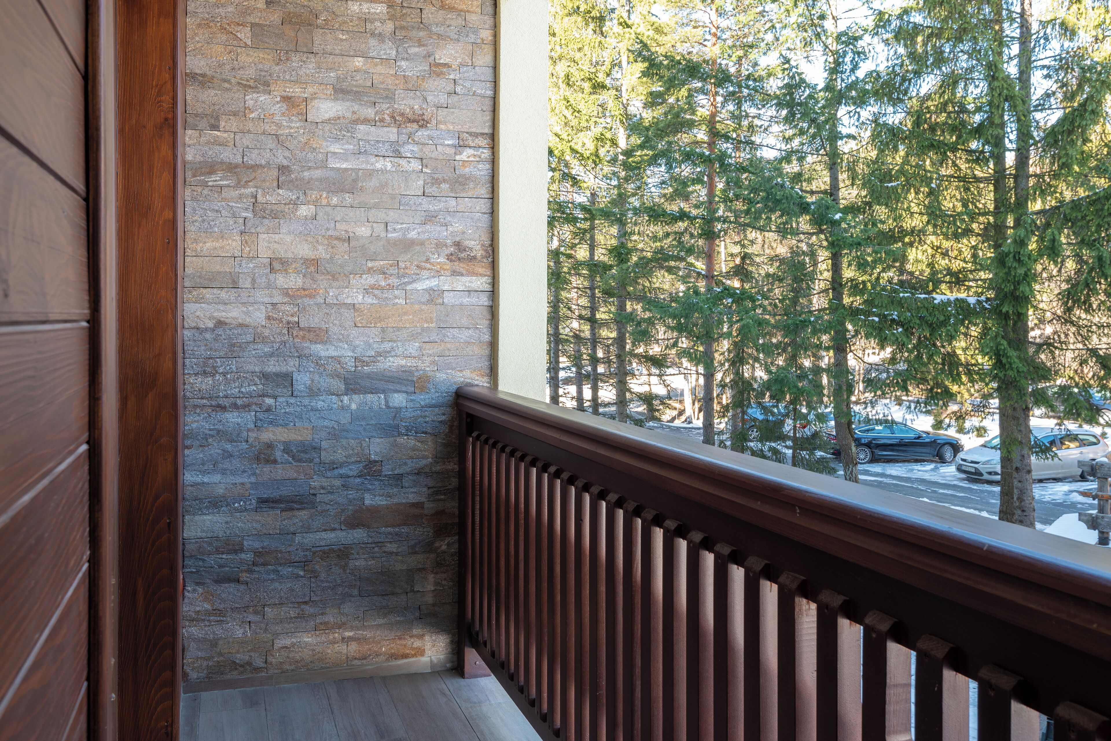 A balcony with a stone wall and a wooden railing, overlooking a street with cars and trees.
