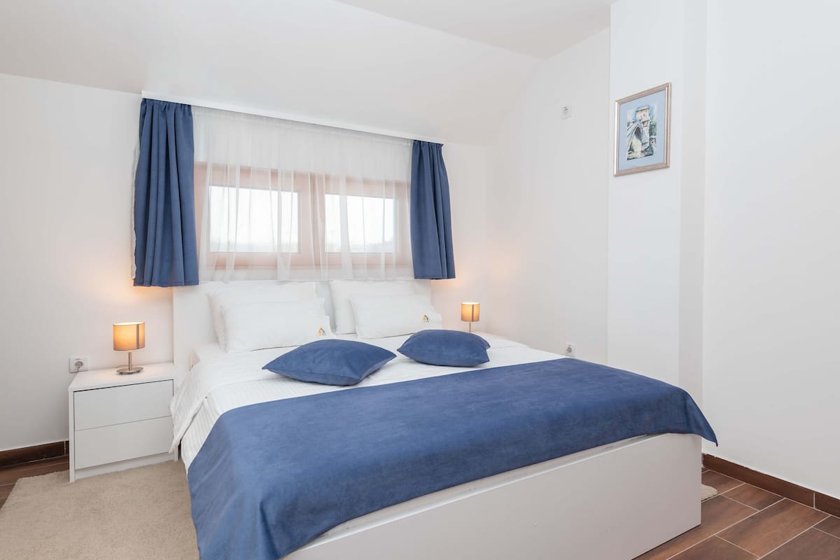 A bedroom with a bed, two bedside tables, a nightstand, a window with sheer curtains, and a framed picture on the wall.