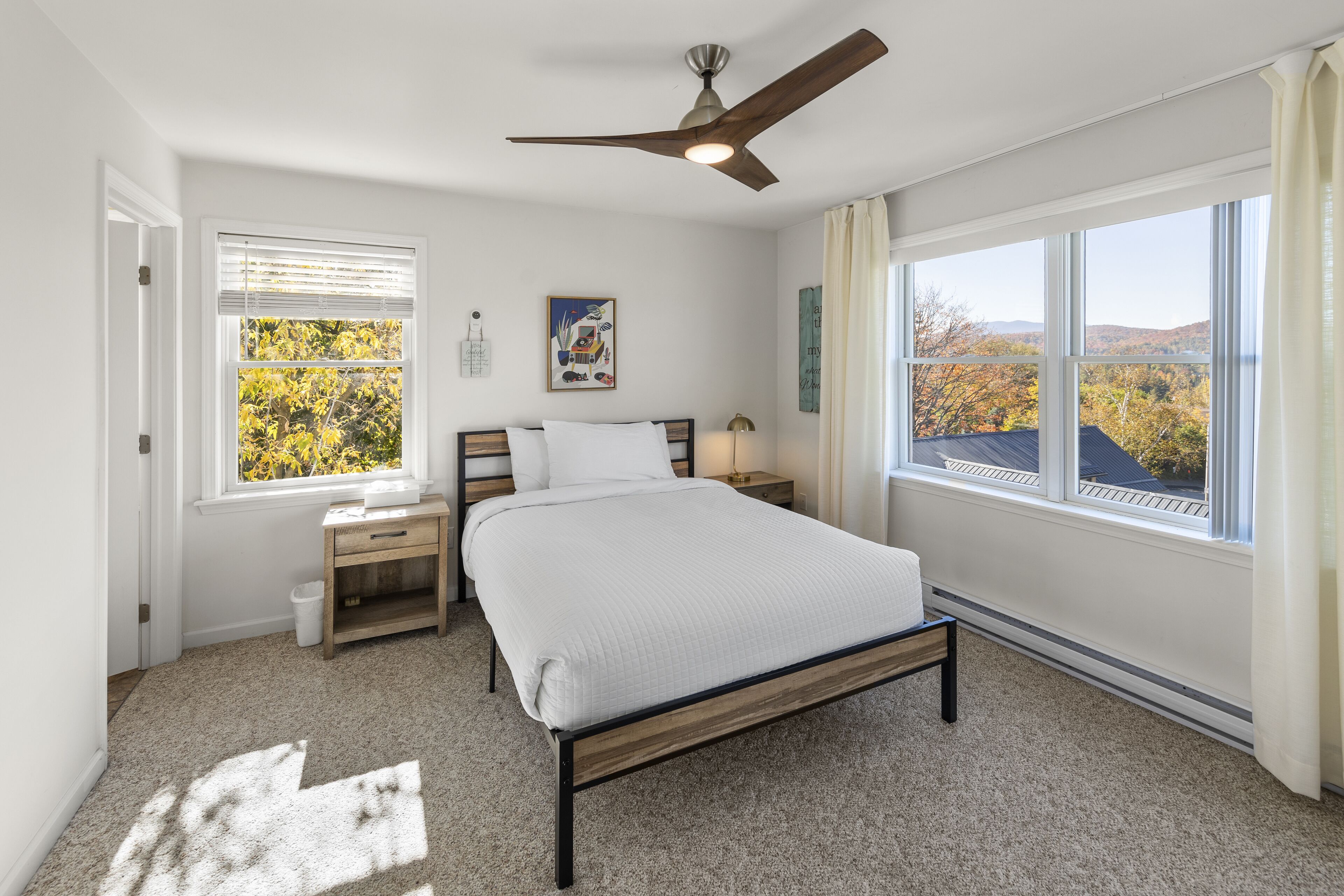 Quarto com cama, mesa de cabeceira, escrivaninha, janela com persianas, ventilador de teto e vista para folhagem outonal.