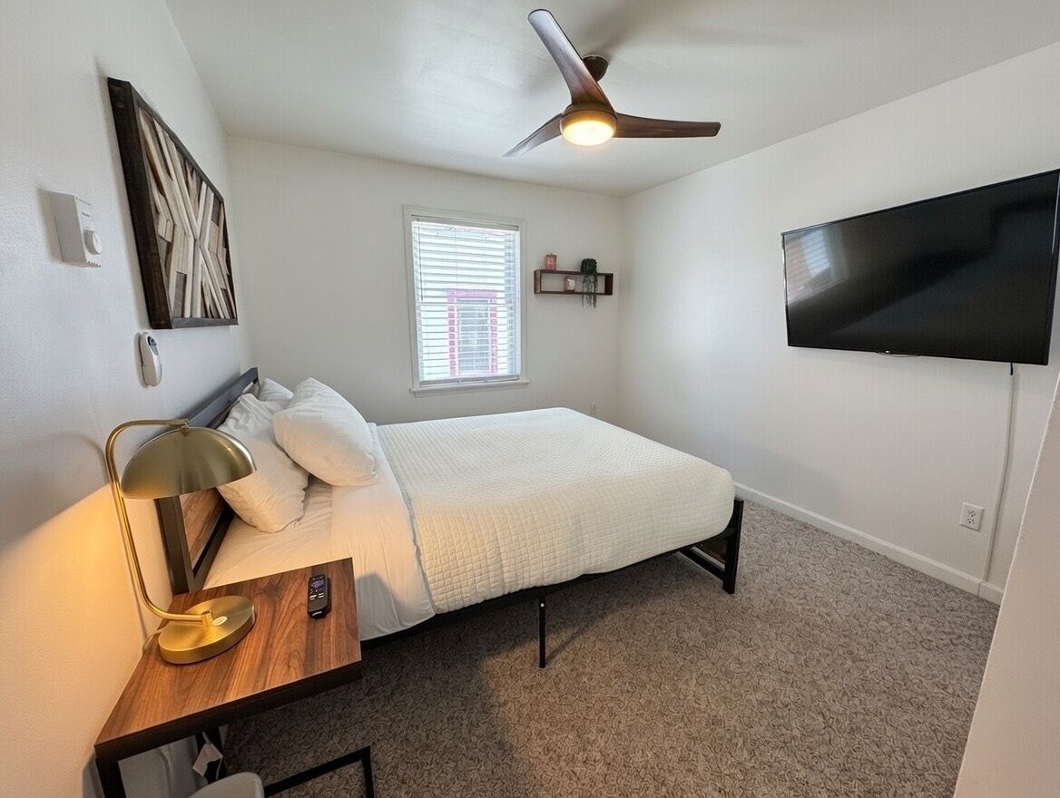 Une chambre à coucher avec un lit, un bureau avec une lampe, un ventilateur de plafond, un téléviseur fixé au mur et une fenêtre avec des stores.