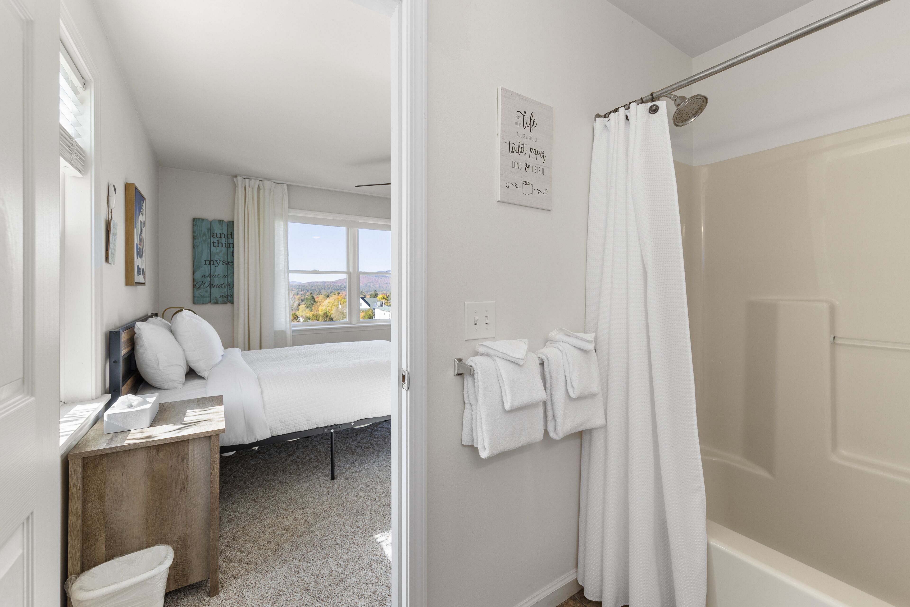 A bedroom with a bed, a wooden bedside table, a window with a view, a shower area, and a wall with a motivational quote.
