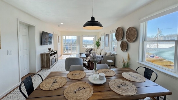 Un comedor con una mesa de madera dispuesta para cuatro personas, rodeada de sillas. Hay individuales tejidos con palabras como “reunirse” y “familia”. Una lámpara colgante negra ilumina la mesa, y la habitación tiene ventanas amplias que ofrecen vistas a un puente y edificios.