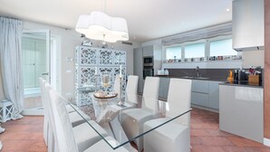A modern kitchen with a central island, a dining area with a glass table and white chairs, and a view of the outside through large windows. - Elegant Villa Rafaelle with pool (Vabriga)