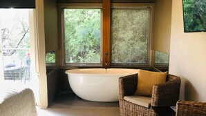 A modern bathroom with a large bathtub, wicker chairs, and a view of greenery through the windows.