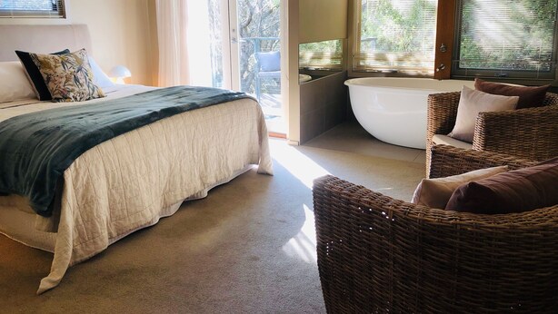 A bedroom with a bed, a wicker chair, a bathtub, and large windows.