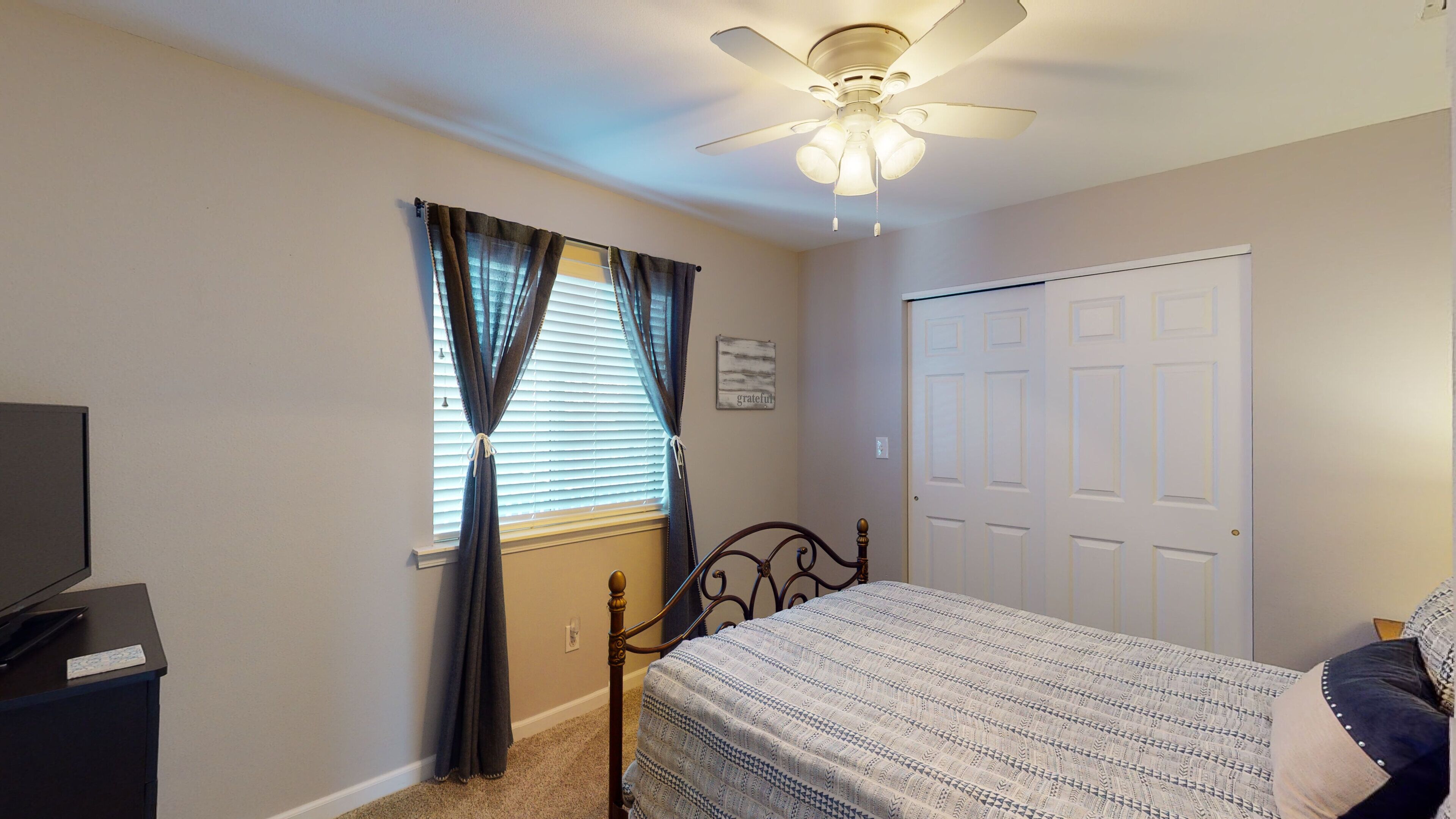 A bedroom with a bed, a ceiling fan, a window with curtains, a door, and a desk with a television.