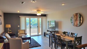 A modern living room with a sofa, chairs, a coffee table, a TV, and a dining area with a table and chairs. - Walk-in Condo! Enjoy The Lake Life, 3B/2B Sleeps 9, 2 Pools, Boat Slip & Beach!  (Osage Beach)