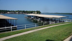 A waterfront property with covered docks and boats, a grassy area, and a clear view of the water. - Walk-in Condo! Enjoy The Lake Life, 3B/2B Sleeps 9, 2 Pools, Boat Slip & Beach!  (Osage Beach)