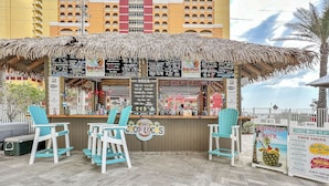 A tiki bar with a thatched roof, multiple seating areas, and a menu board listing various drinks and food items.