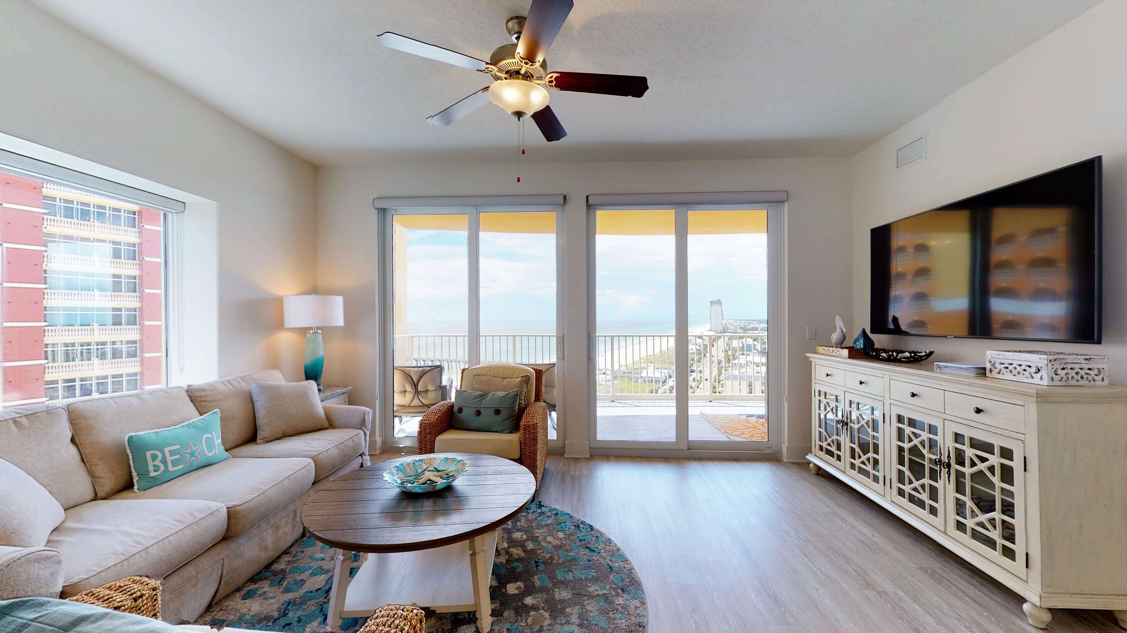 A living room with a sofa, coffee table, and a TV on a cabinet. There is a balcony with a view of the city.