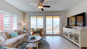 A living room with a sofa, coffee table, and a TV on a cabinet. There is a balcony with a view of the city.