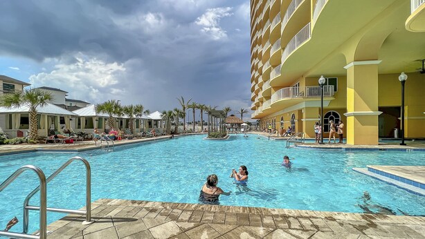 A large outdoor swimming pool surrounded by palm trees and a multi-story hotel building.