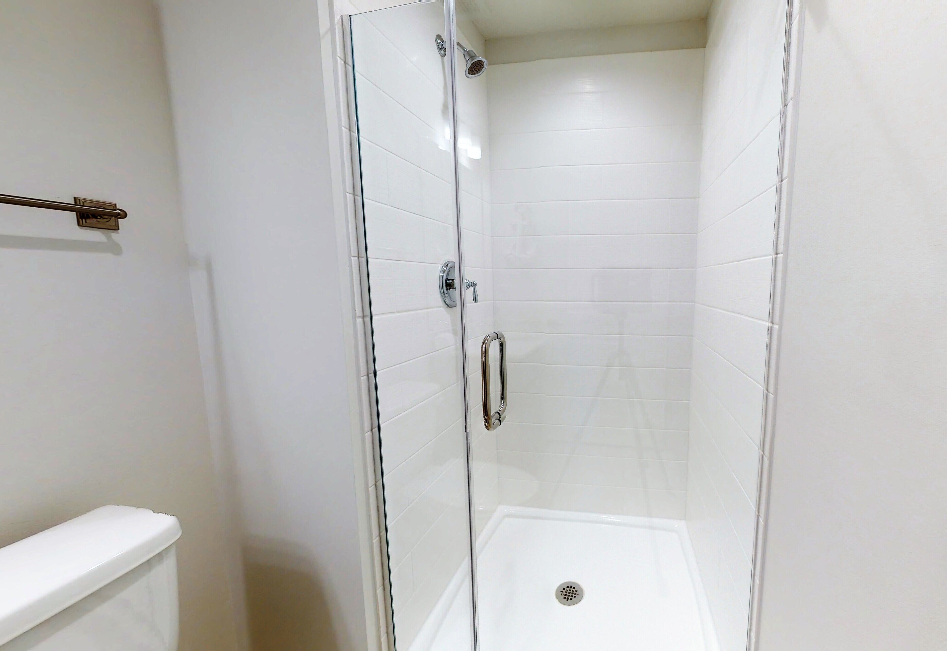 Un vestuario de ducha con paredes y piso de azulejos blancos, desagüe visible y un perchero metálico en la pared.