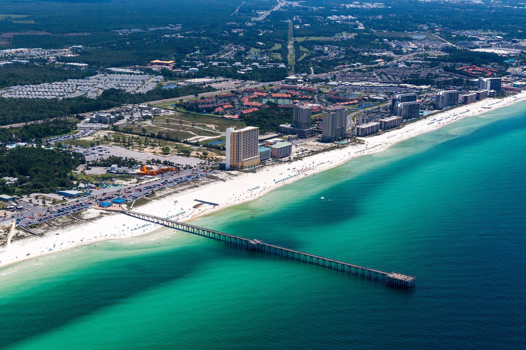 Una vista aerea di una città costiera con un lungo molo che si protende verso il mare, circondata da una spiaggia, hotel e una zona urbana densa.