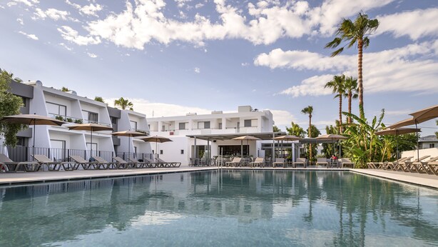 Una piscina con sillas de descanso y sombrillas, rodeada por un edificio moderno con balcones y palmeras.