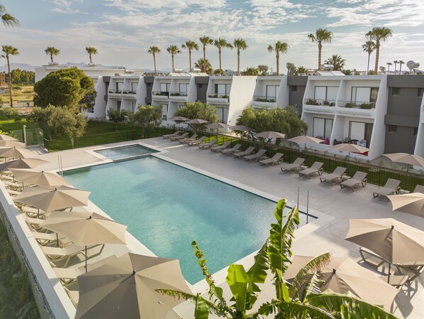 A swimming pool with lounge chairs and umbrellas, surrounded by palm trees and modern buildings. - Smaragdi Kos Hotel (Kos)